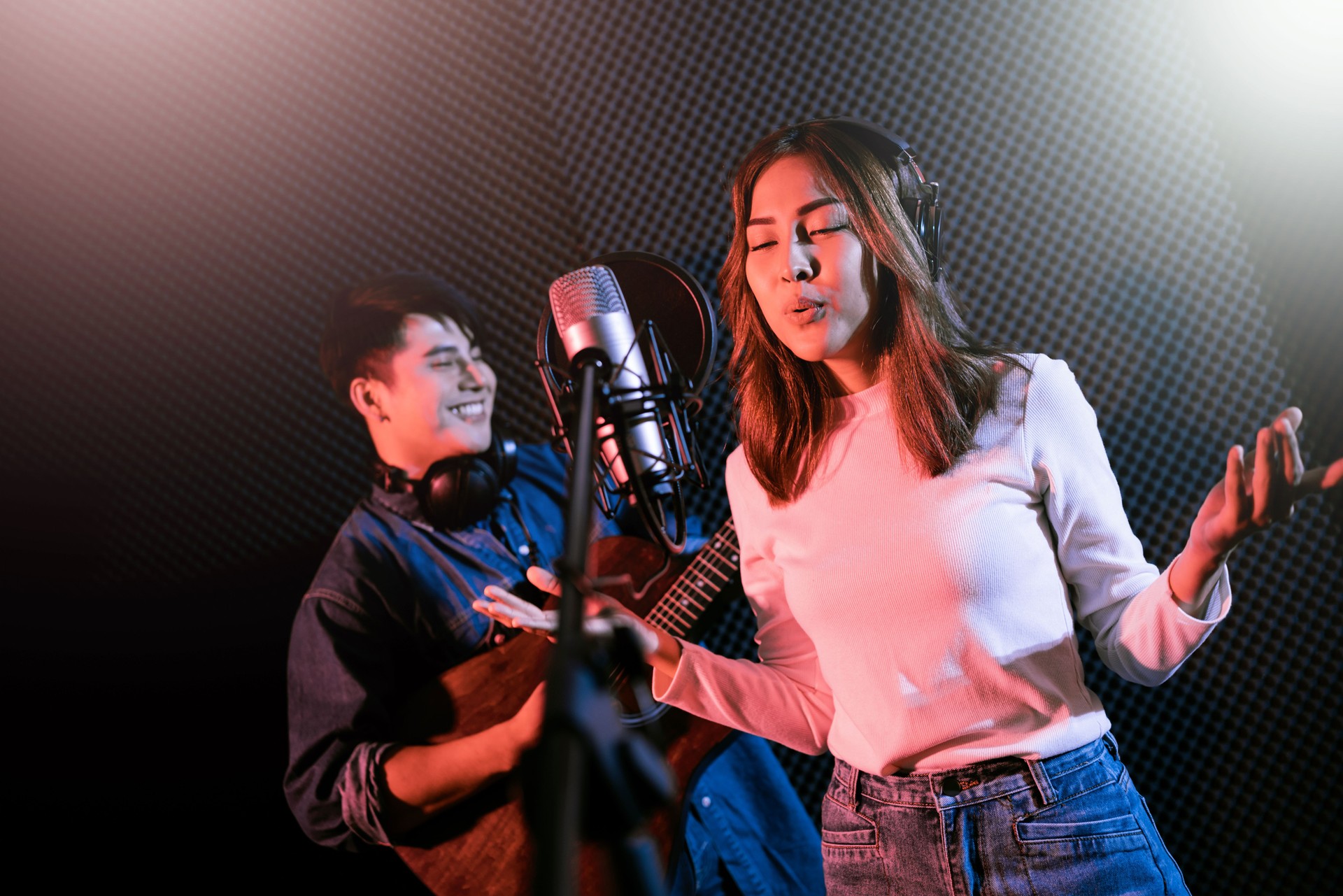 Hübsche asiatische Sängerin, die Songs mit einem Studiomikrofon und einem Pop-Schild am Mikrofon aufnimmt, wobei der Mann Gitarre in blauem und rotem Licht spielt. Performance und Show im Musikbusiness. Duett-Sitzung.